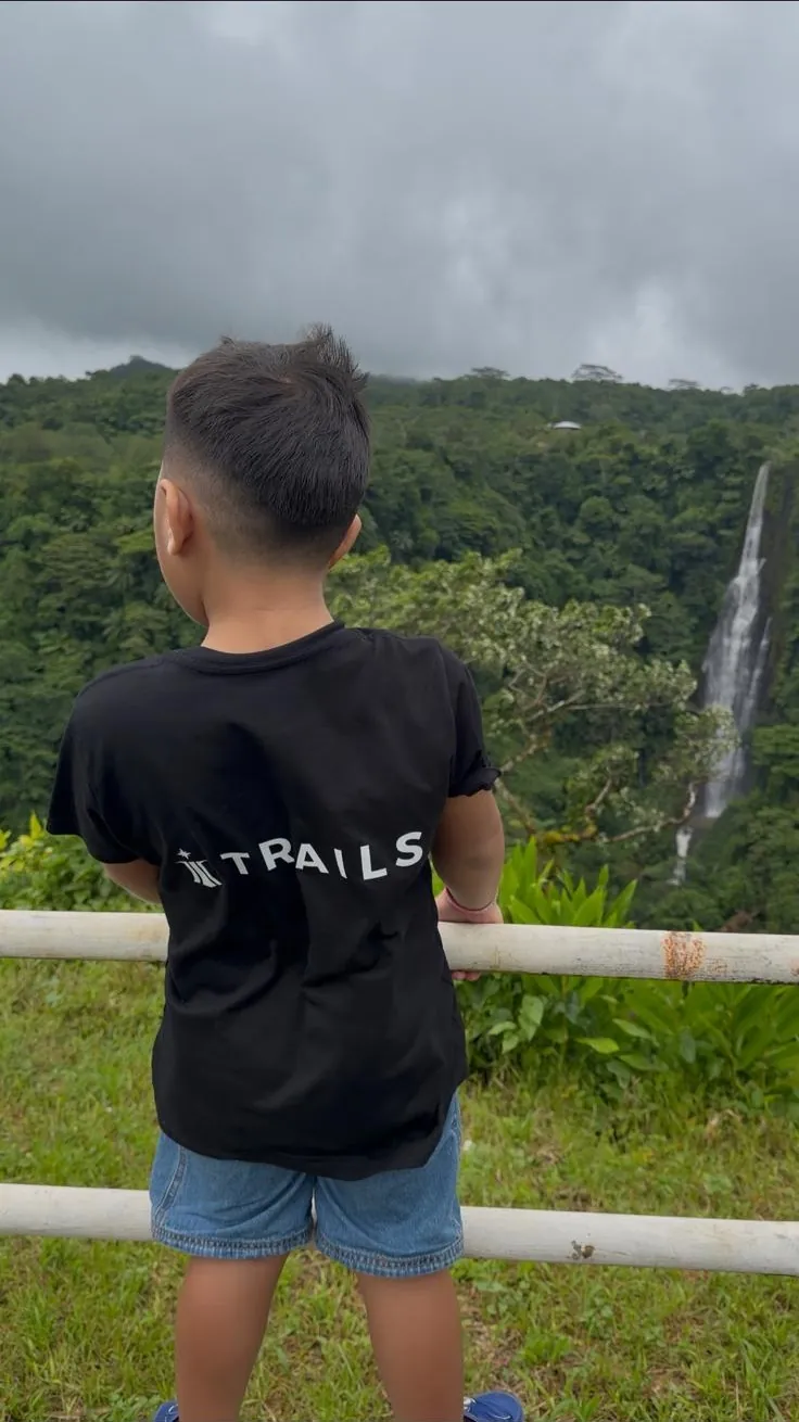 A child in a TRAILS t-shirt looking out over a rainforest valley and waterfall in Samoa